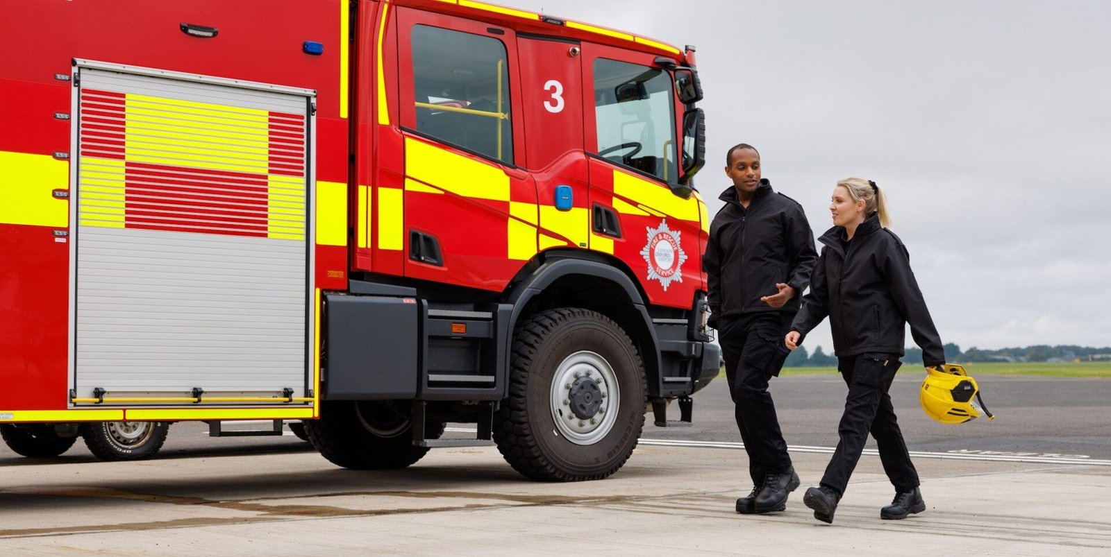 Fire Crew wearing HeathBrook jackets and trousers - black. Male and female fire fighter by fire engine just off the runway.